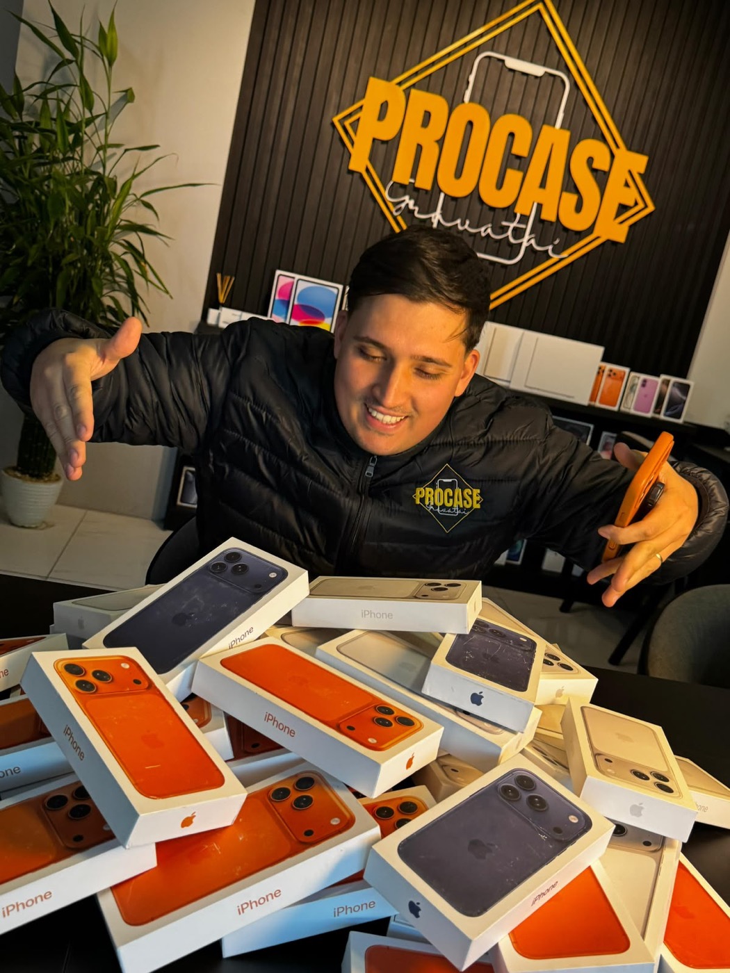 Juliano, fundador da Procase, debruçado sobre uma pilha de caixas de iPhone, com o letreiro da loja ao fundo