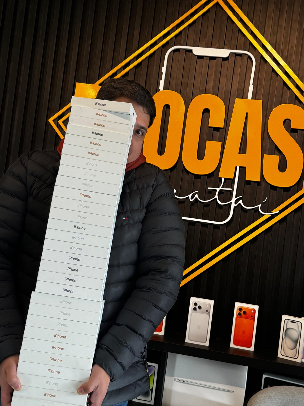 Juliano, fundador da Procase, segurando uma pilha de caixas de iPhone diante da prateleira de produtos da loja